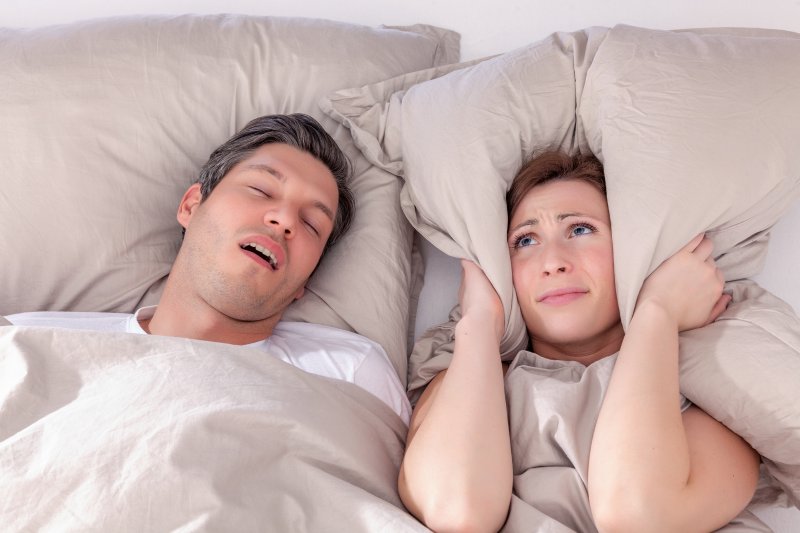 a young couple lying in bed with the man snoring and the woman covering her ears with a pillow 